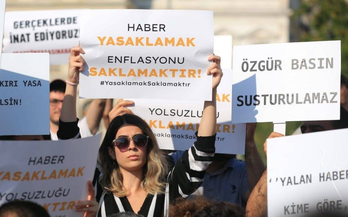 Görüntüde kalabalık bir protesto sahnesi var. Ön planda, güneş gözlüğü takan bir kadın iki eliyle başının üzerinde bir pankart tutuyor. Pankartta büyük harflerle “HABER YASAKLAMAK ENFLASYONU SAKLAMAKTIR!” yazıyor ve altında “#YasaklamakSaklamaktır” etiketi yer alıyor.