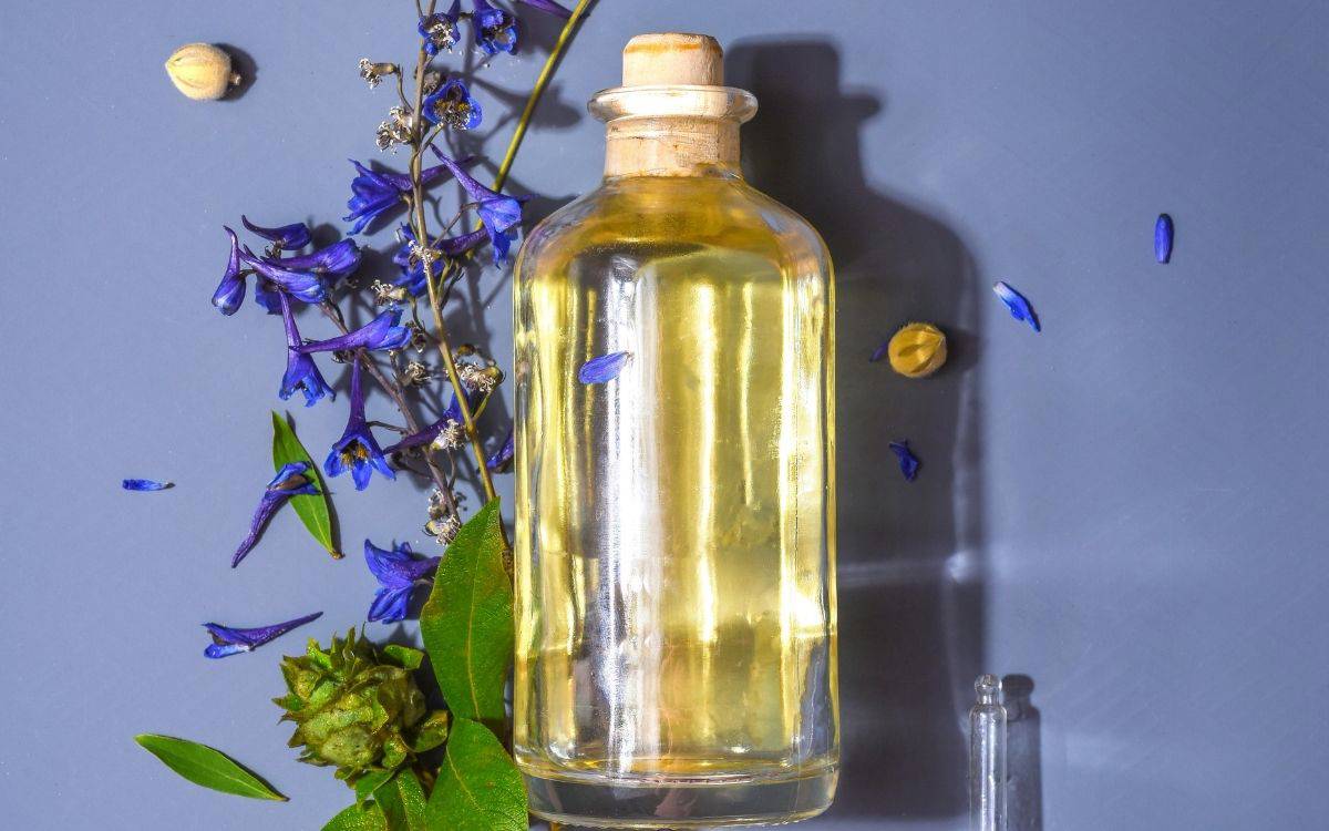 Mavi zeminde mantarlı cam şişede açık sarı yağ duruyor; çevresine mor çiçekler, yeşil yapraklar ve birkaç küçük tohum serpiştirilmiş.