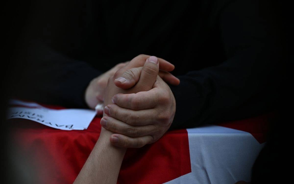 Fotoğrafta karanlık bir fonda, ellerini sıkıca kenetlemiş bir kişi görülüyor. Kişinin üstünde siyah kıyafet var; yüzü görünmüyor, kadraj yalnızca ellerine ve kısmen kollarına odaklanıyor. Ellerin altında kırmızı-beyaz bir örtü ya da bayrak benzeri bir kumaş yer alıyor. Sol tarafta, kumaşın üstünde kısmen görünen beyaz bir kâğıt da var. Genel atmosferi sessiz, ağır ve yas duygusu taşıyan bir sahne gibi görünüyor.