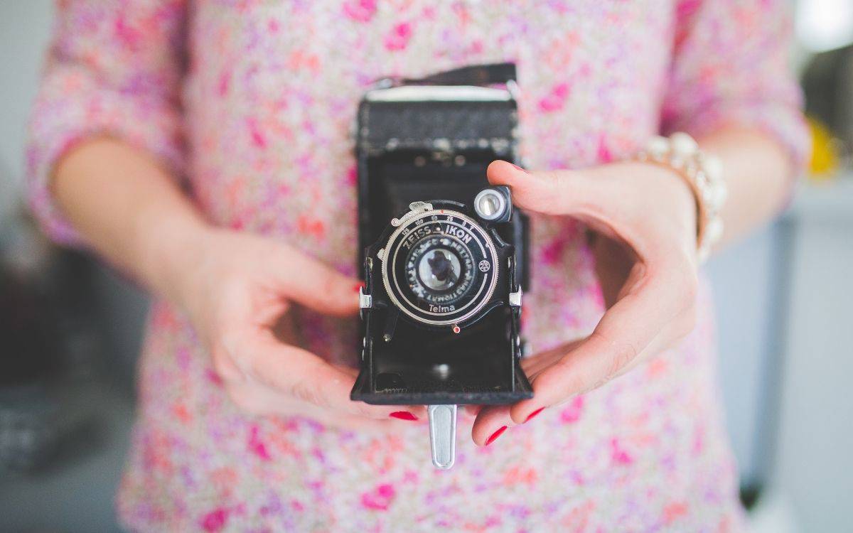 Pembe çiçekli kıyafet giyen bir kişinin elleri, objektifi öne dönük eski bir fotoğraf makinesini yakın planda tutuyor.