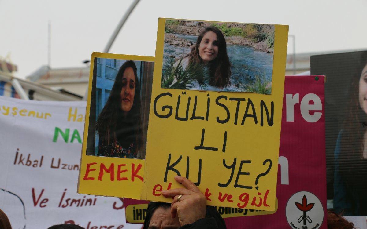 Kalabalık bir eylemde havaya kaldırılan sarı dövizde iki genç kadının fotoğrafı ve büyük siyah harflerle yazılmış bir slogan var; arka planda başka pankartlar görünüyor.