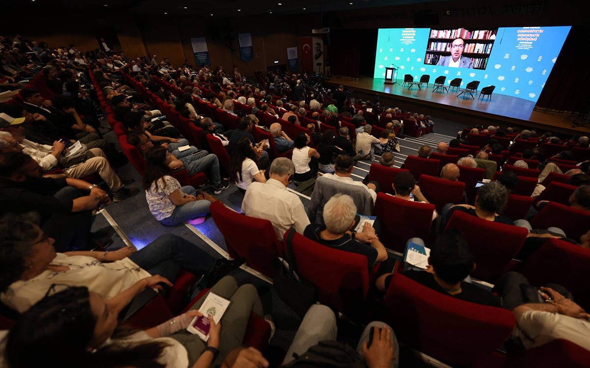 Geniş bir konferans salonunda çok sayıda katılımcı kırmızı koltuklarda oturuyor. Sahnedeki büyük ekranda çevrim içi bağlanan bir konuşmacı görülüyor; ekranın yanında konferansın “Cumhuriyetin Demokratik Dönüşümü” başlıklı görselleri yer alıyor. Sahnenin önünde boş sandalyeler, salonda ise etkinliği takip eden kalabalık bir izleyici grubu bulunuyor.